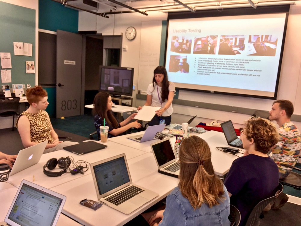 Presenting usability testing findings to a team seated around laptops in NYU's Ability Studio. The slide lists issues found on the app and website, including content linearization, missing feedback on interactions, insufficient labelling of buttons and input fields, timeouts, pinch and zoom being disabled, and touch gestures screen reader users rely on not being available on tablet.